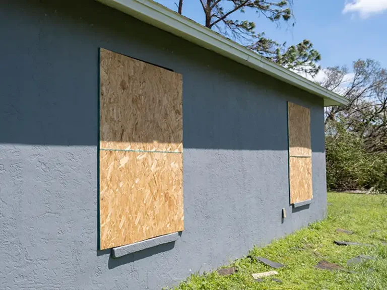 Windows boarded up with plywood on a gray house exterior.