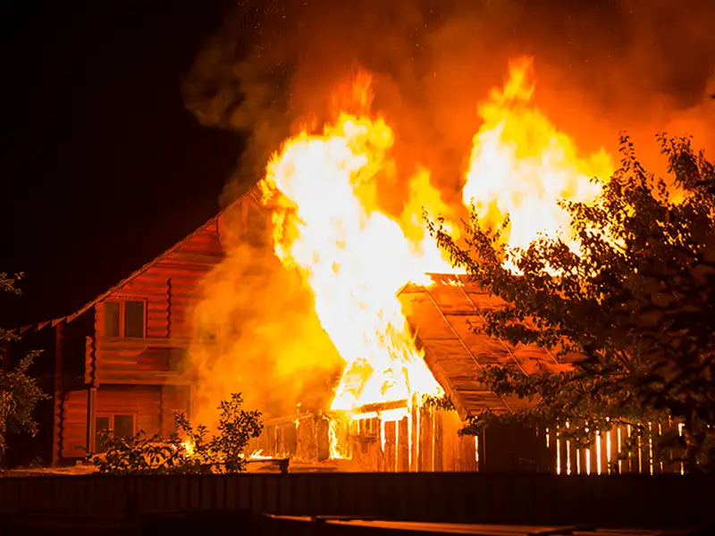Wooden house engulfed in large flames at night with smoke rising.