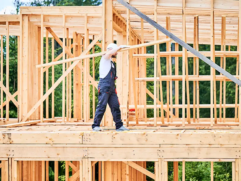 Construction worker carrying a wooden plank on a house frame under construction.