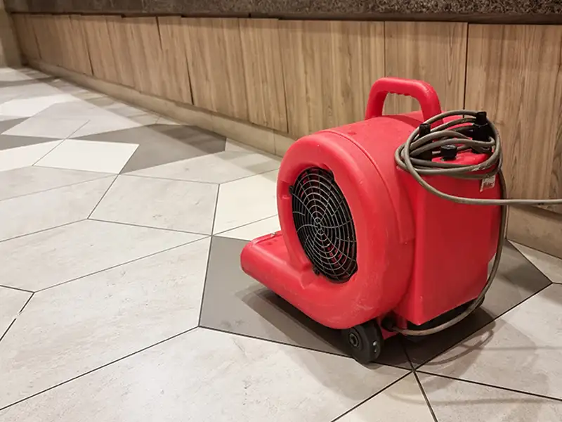 Red industrial air blower with a coiled power cord on a tiled floor near a wooden wall.