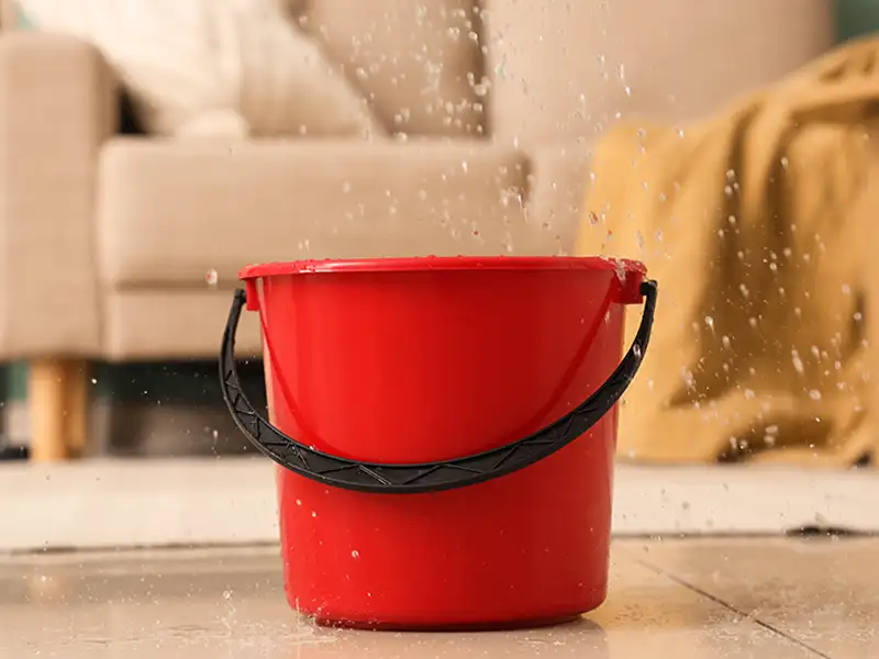Red plastic bucket catching water drops on a tiled floor indoors.